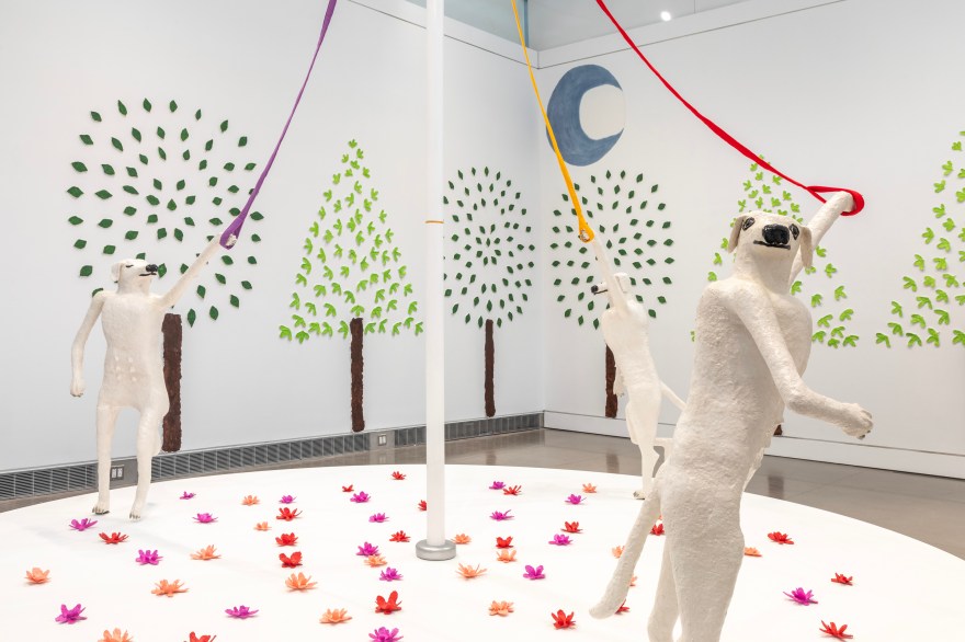 Three white dogs stand upright around a white cane maypole. On the right, a dog holding a red leash looks over her shoulder right at us. They're among a field of red, pink, and magenta flowers, and a field of light and dark green trees. Behind them is a grey-blue crescent moon.