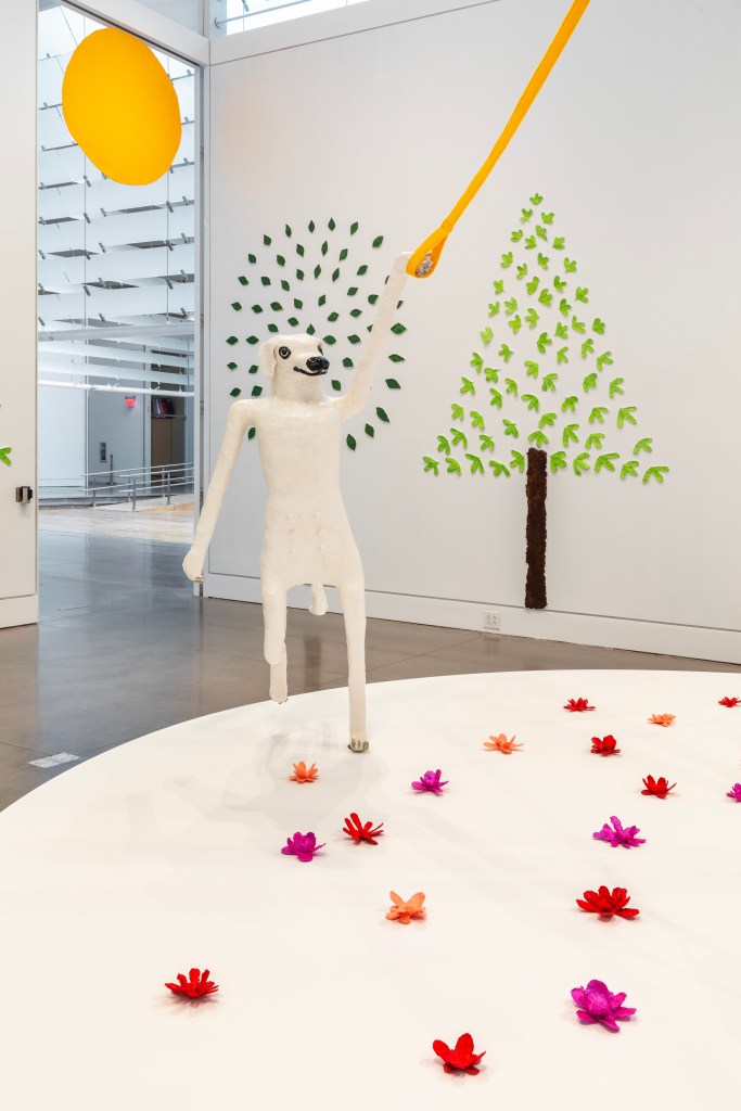 Sculpture of white dog standing on her hind legs, walking towards us while holding onto a yellow ribbon. She stands among a field of flowers and trees, and has a bright yellow sun above her