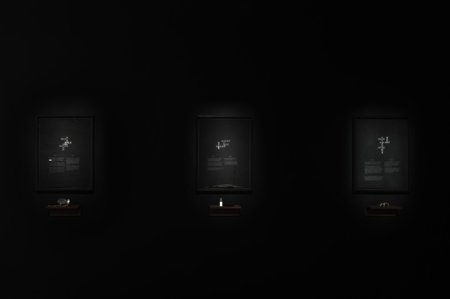 Install view of 3 black works on paper dramatically lit against a black gallery wall. The 3 pieces feature silvery crossword puzzles. Below each is a walnut shelf featuring knicknacks.