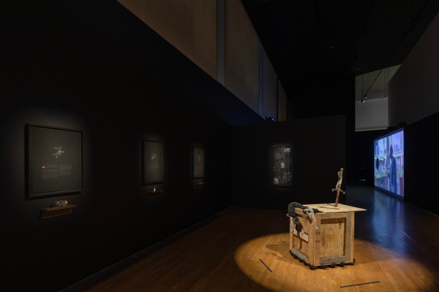 Dimly lit gallery with black walls features dramatically lit artworks, including 4 vertically hung black and white works on paper, 1 box-like sculpture, and 1 large film screening