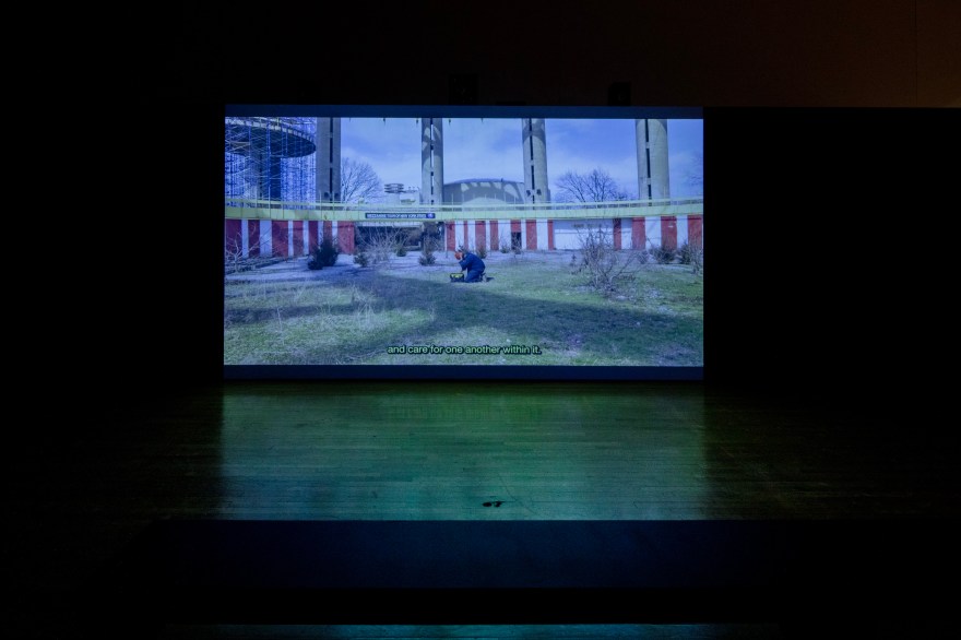 Film screening in dimly lit gallery. On screen is a Black man, kneeling within an enclosed green field. Surrounding him is red and white striped circus-like architecture.