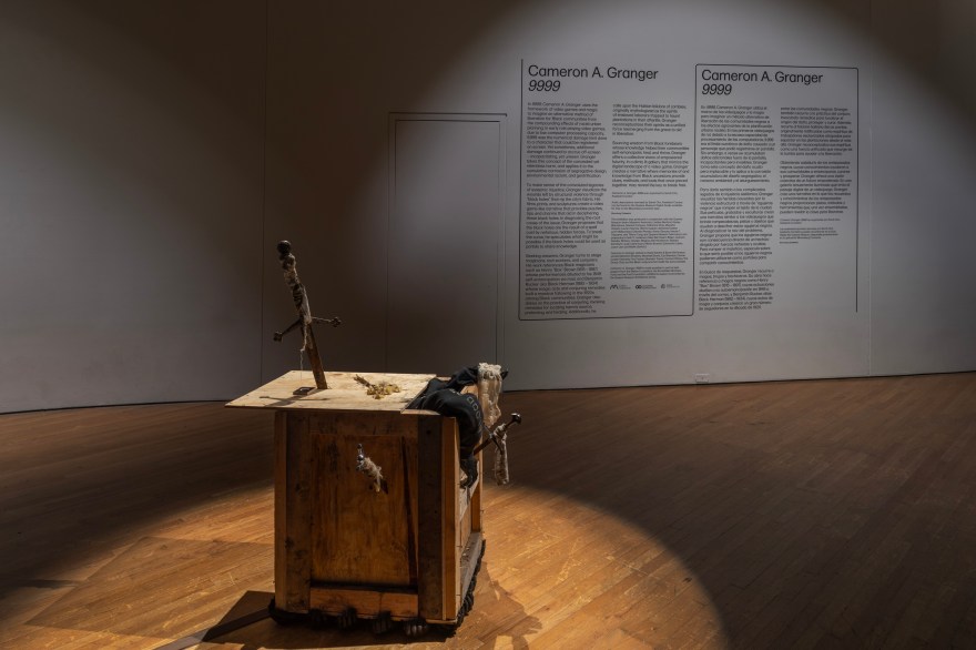 a horizontal view of a crate box sculpture with a black figure emerging. The sculpture is brightly lit in a dim gallery. Behind it on a white wall is the black vinyl exhibition wall text about the show. It's 4 columns long.